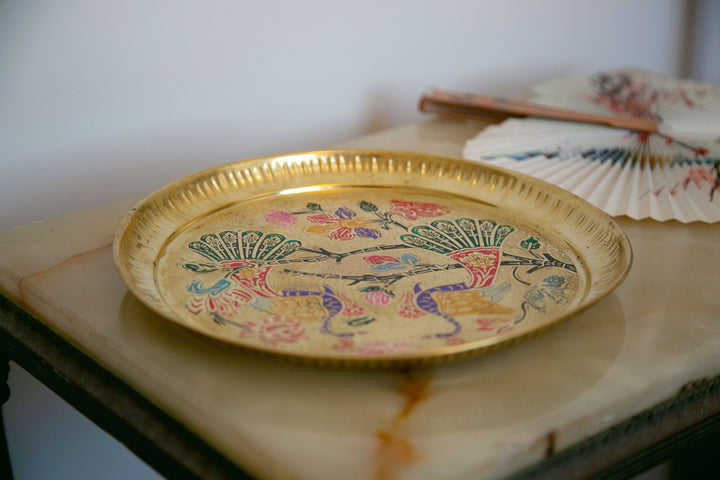 Golden decorative plate with colorful bird and floral designs, placed on marble with a patterned fan in the background.
