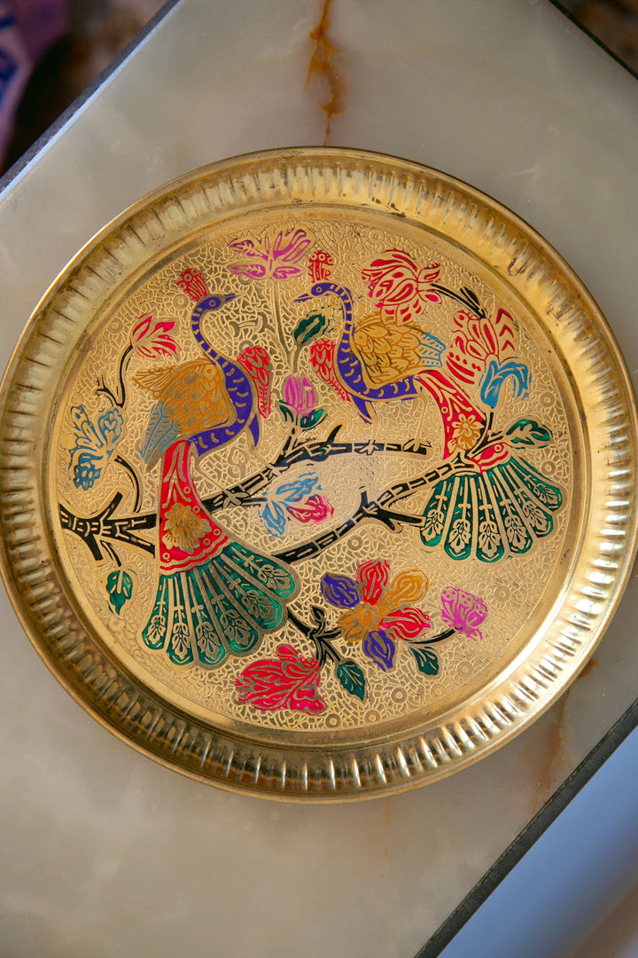 A golden metal tray with a peacock and floral designed centerpiece.