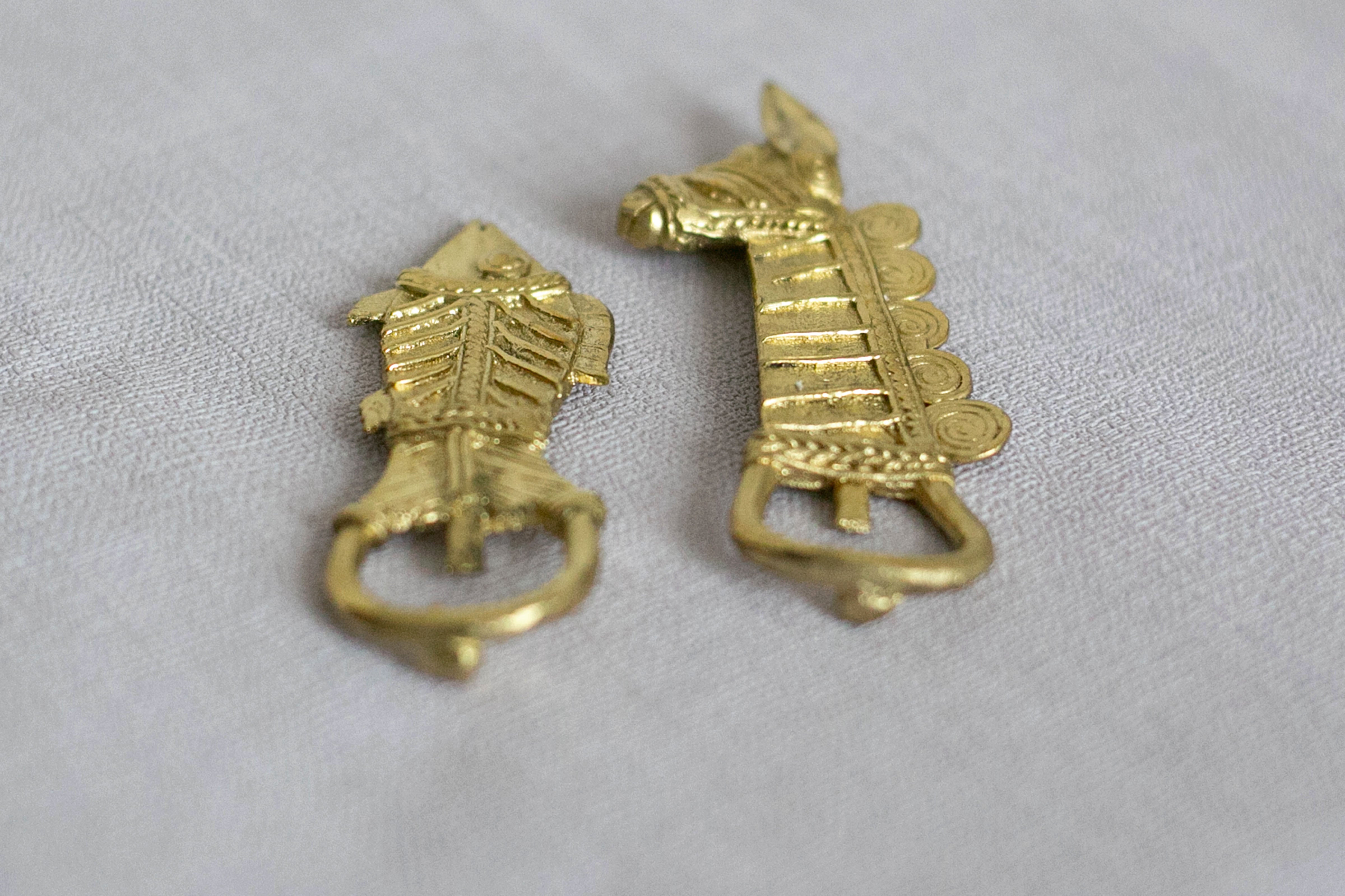 A set of two gold-colored brass metal bottle openers with horse and fish designs, placed on a white fabric surface.