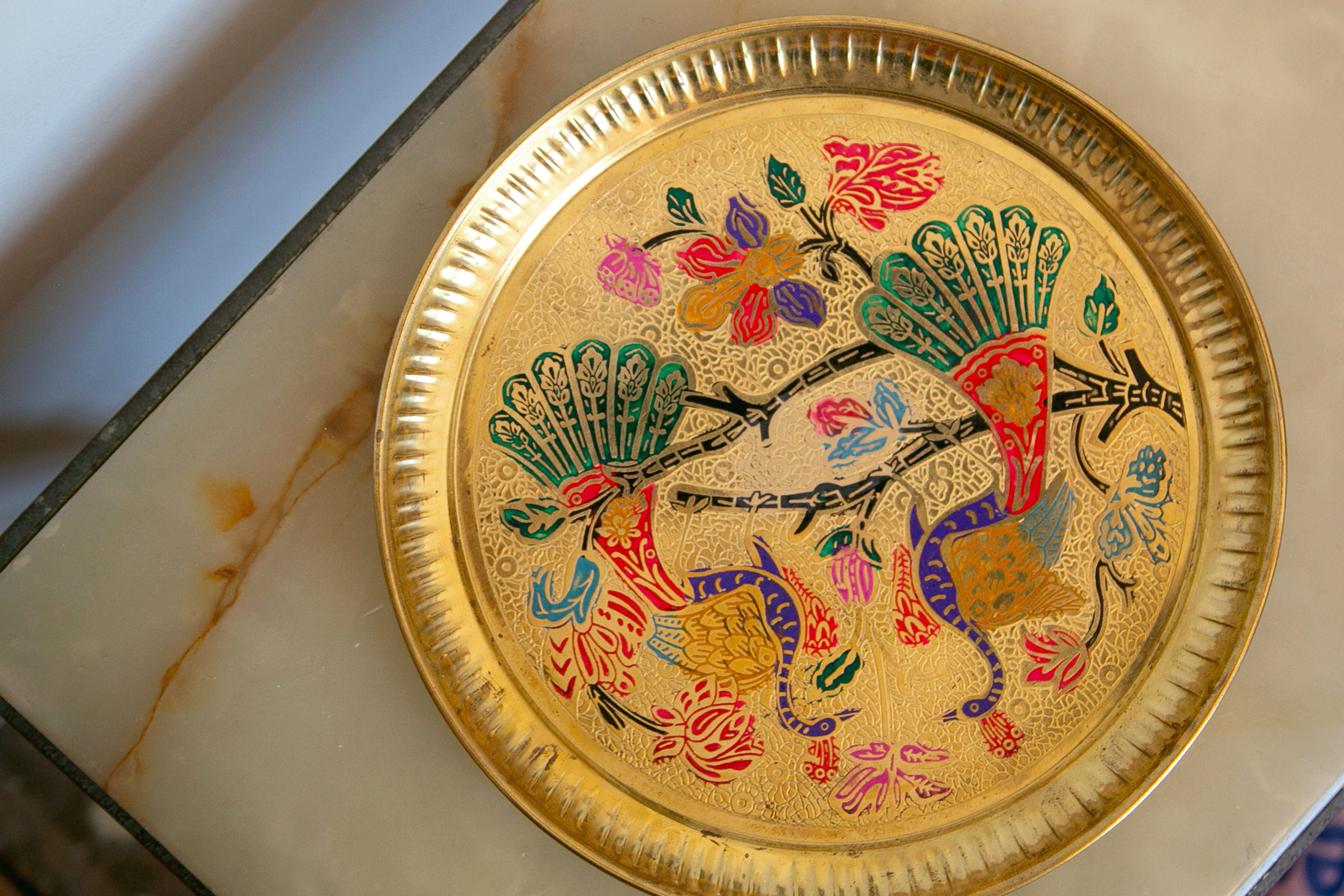 Decorative golden plate featuring two stylized peacocks with colorful tail feathers, surrounded by intricate floral patterns, resting on a marble surface.