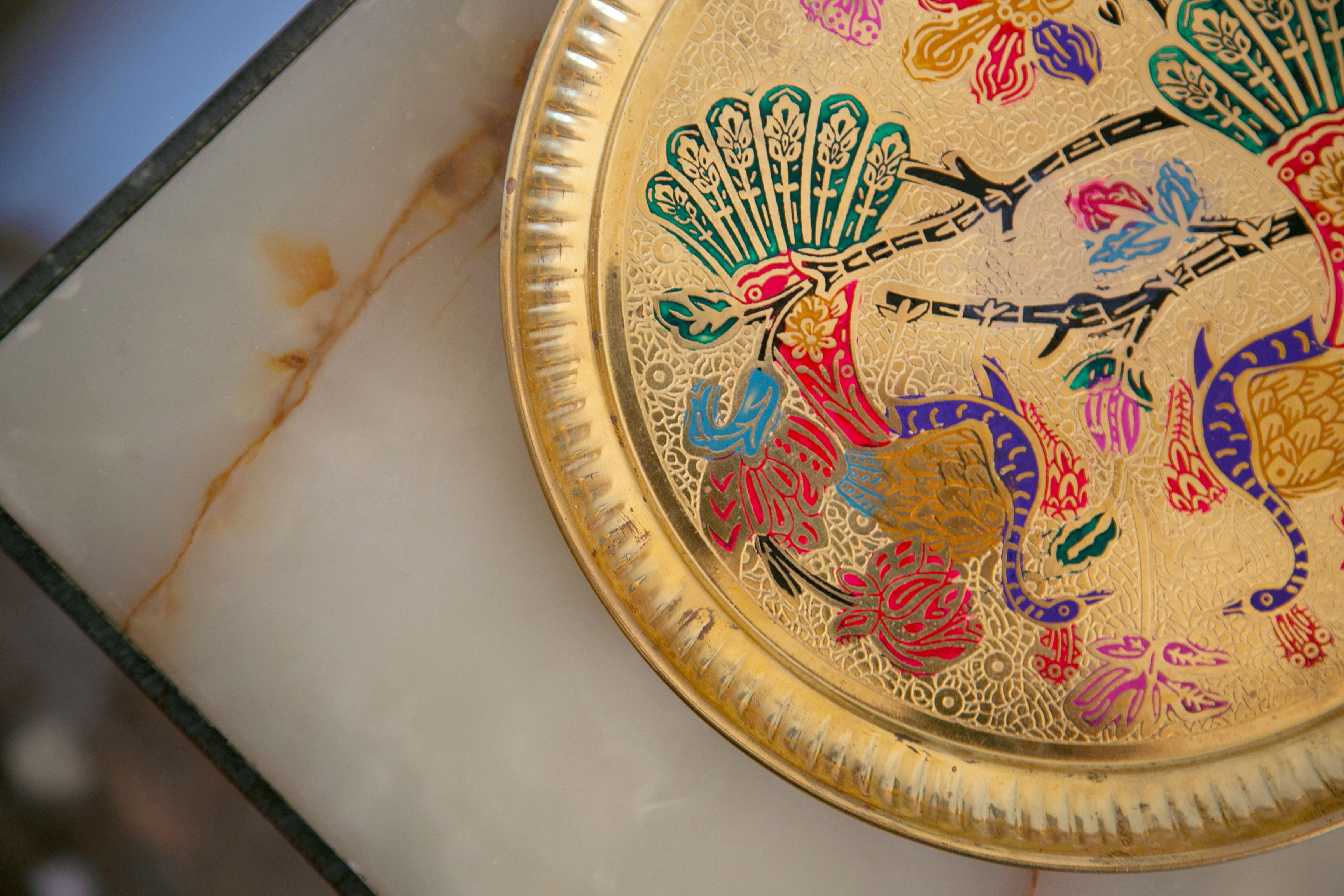 Decorative golden plate with colorful bird and floral designs resting on a marble surface.