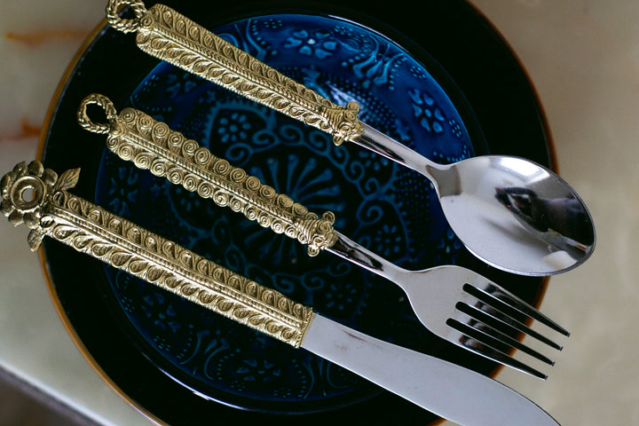 A set of dining flatware consisting of a fork, knife, and spoon with ornate gold-plated handles, displayed on a blue patterned plate.
