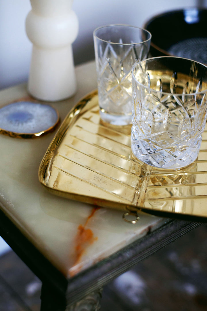 Two clear glasses on a gold-colored serving tray with a geometric pattern, placed on a marble surface with a decorative object in the background.