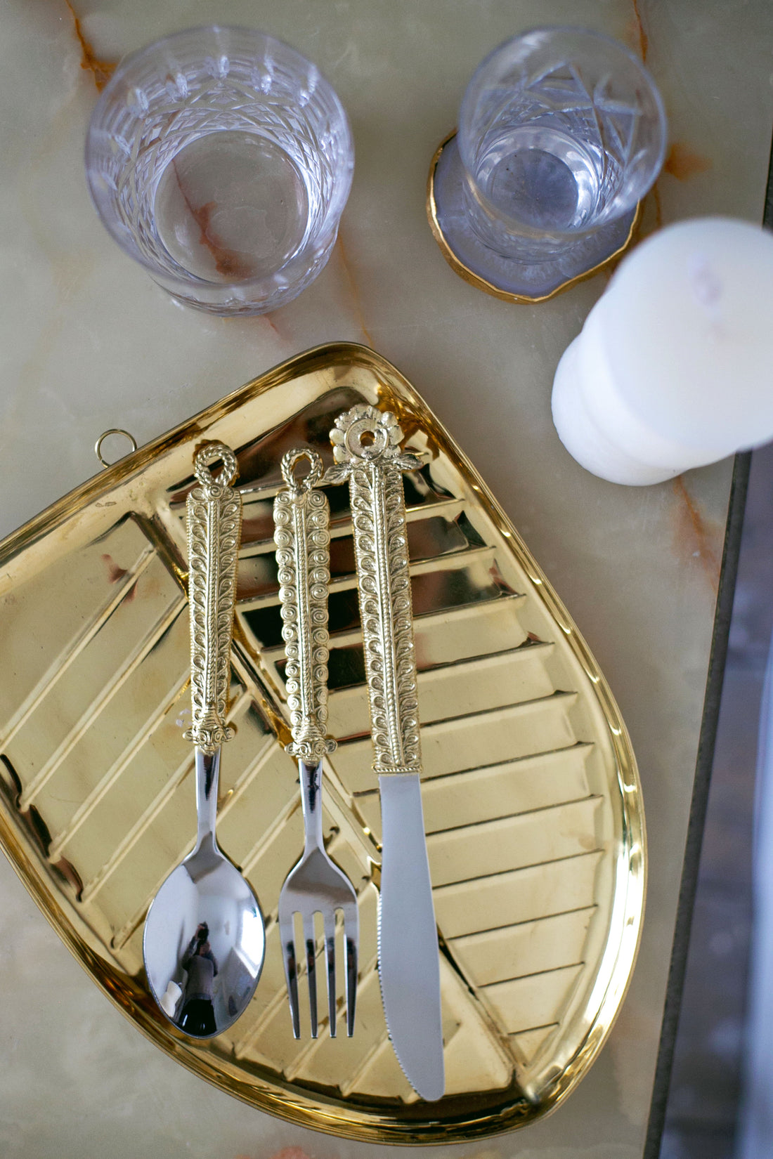 Gold tray with ornate spoon, fork, and knife, alongside two clear textured glasses on a marble surface.
