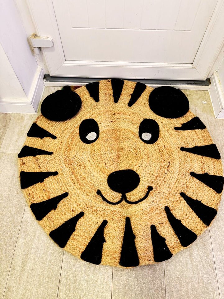 A beige and black jute braided rug with a design resembling a lion's face, placed on a floor with an open door in the background.