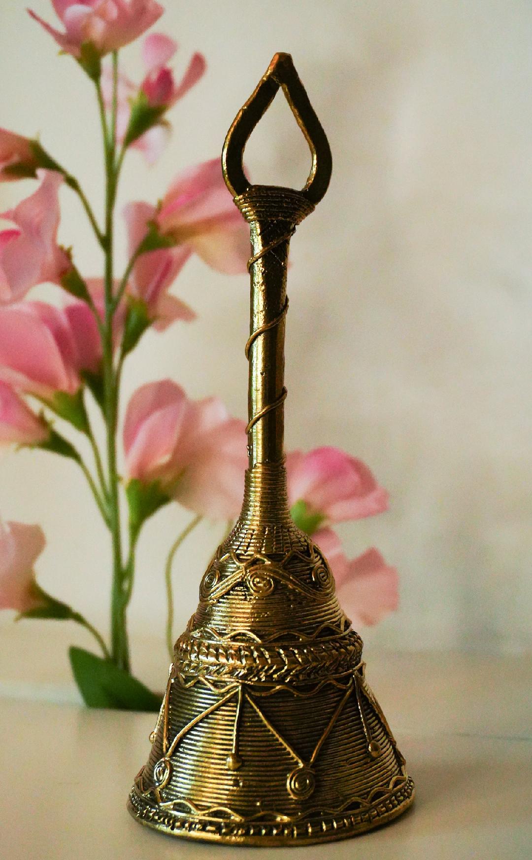A handcrafted brass metal handbell with intricate patterns, displayed against a floral background.