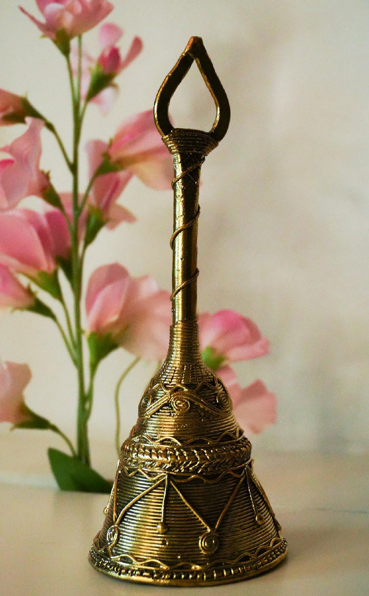 A handcrafted brass metal handbell with intricate patterns, displayed against a floral background.
