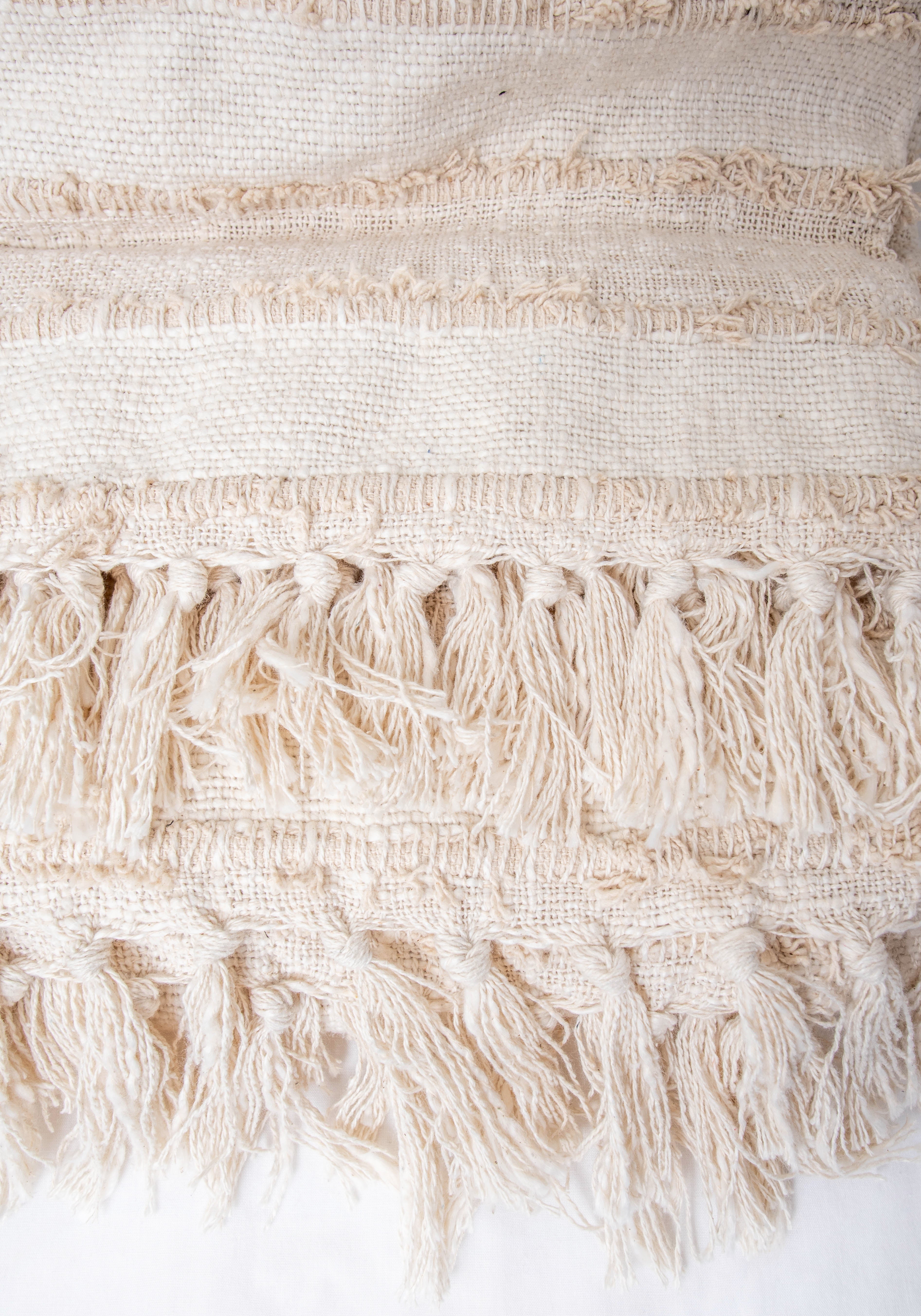 This beige cotton throw blanket features a sophisticated striped pattern, fringed edges, and a textured weave. 