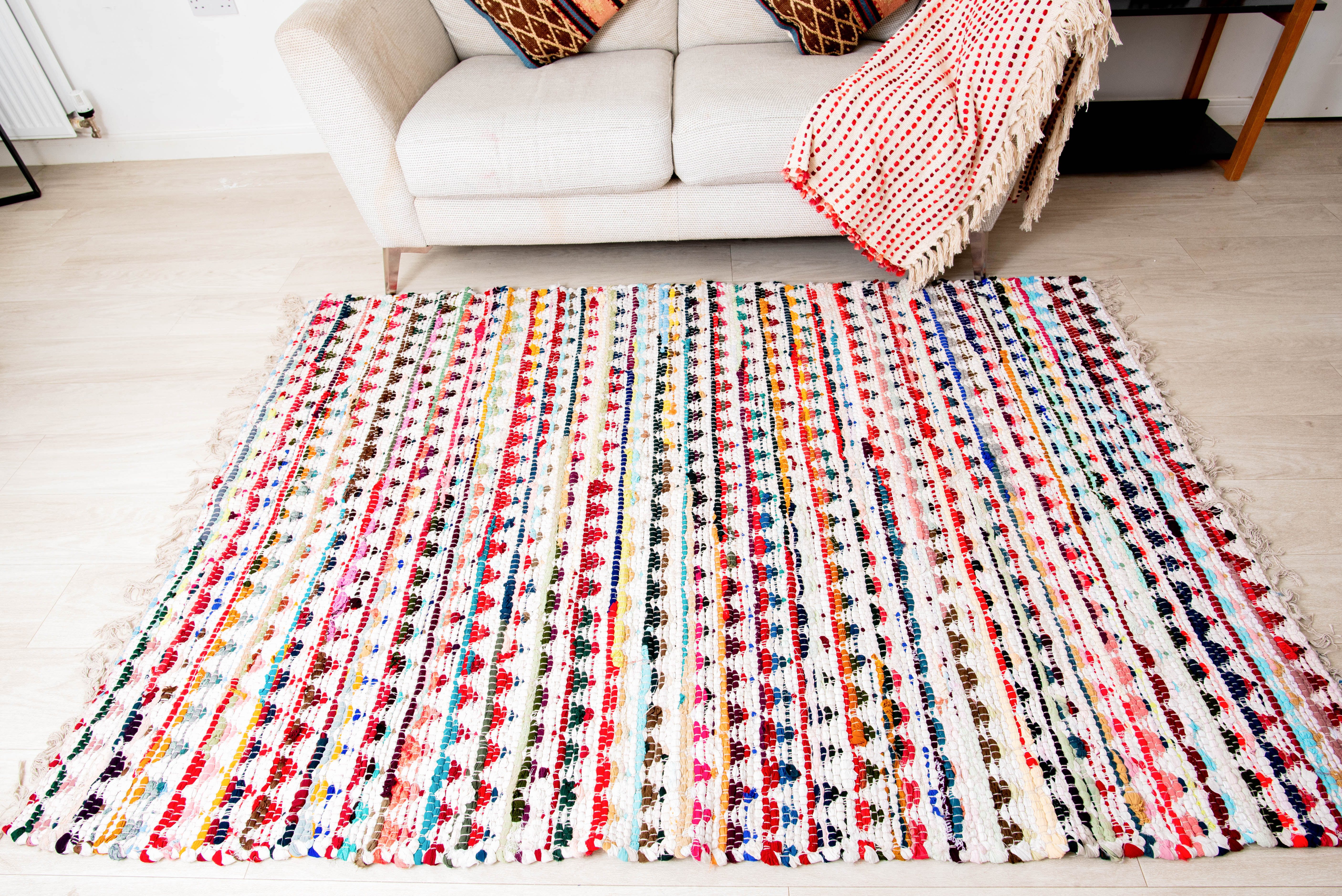 A multicolored geometric patterned cotton rug placed on a floor in a living room setting, with a white sofa and a red striped chair in the background.