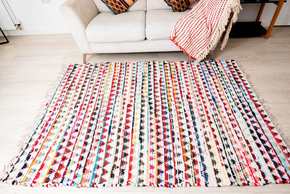 A multicolored geometric patterned cotton rug placed on a floor in a living room setting, with a white sofa and a red striped chair in the background.