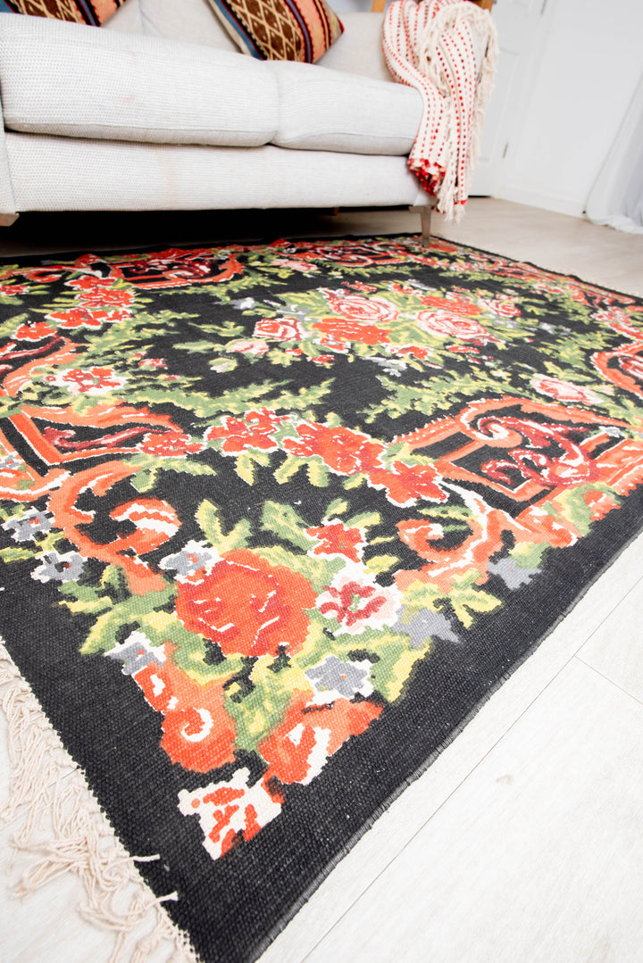 A rectangular, floral design cotton rug in shades of black, red, and green, placed on a floor with a white door in the background.