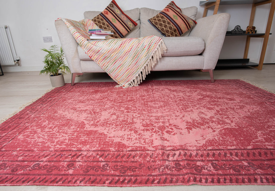 A rectangular, traditional red braided rug with floral patterns, placed on a living room floor next to a sofa with colorful cushions and a plant.