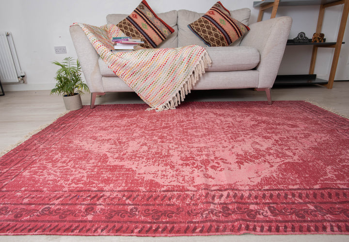 A rectangular, traditional red braided rug with floral patterns, placed on a living room floor next to a sofa with colorful cushions and a plant.