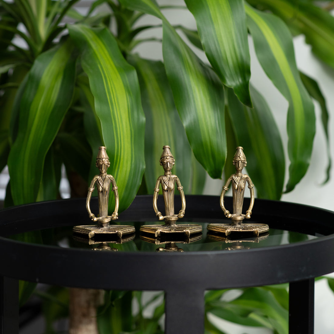 Three brass metal sculptures of sitting women musicians on a round black table, with a green plant in the background.