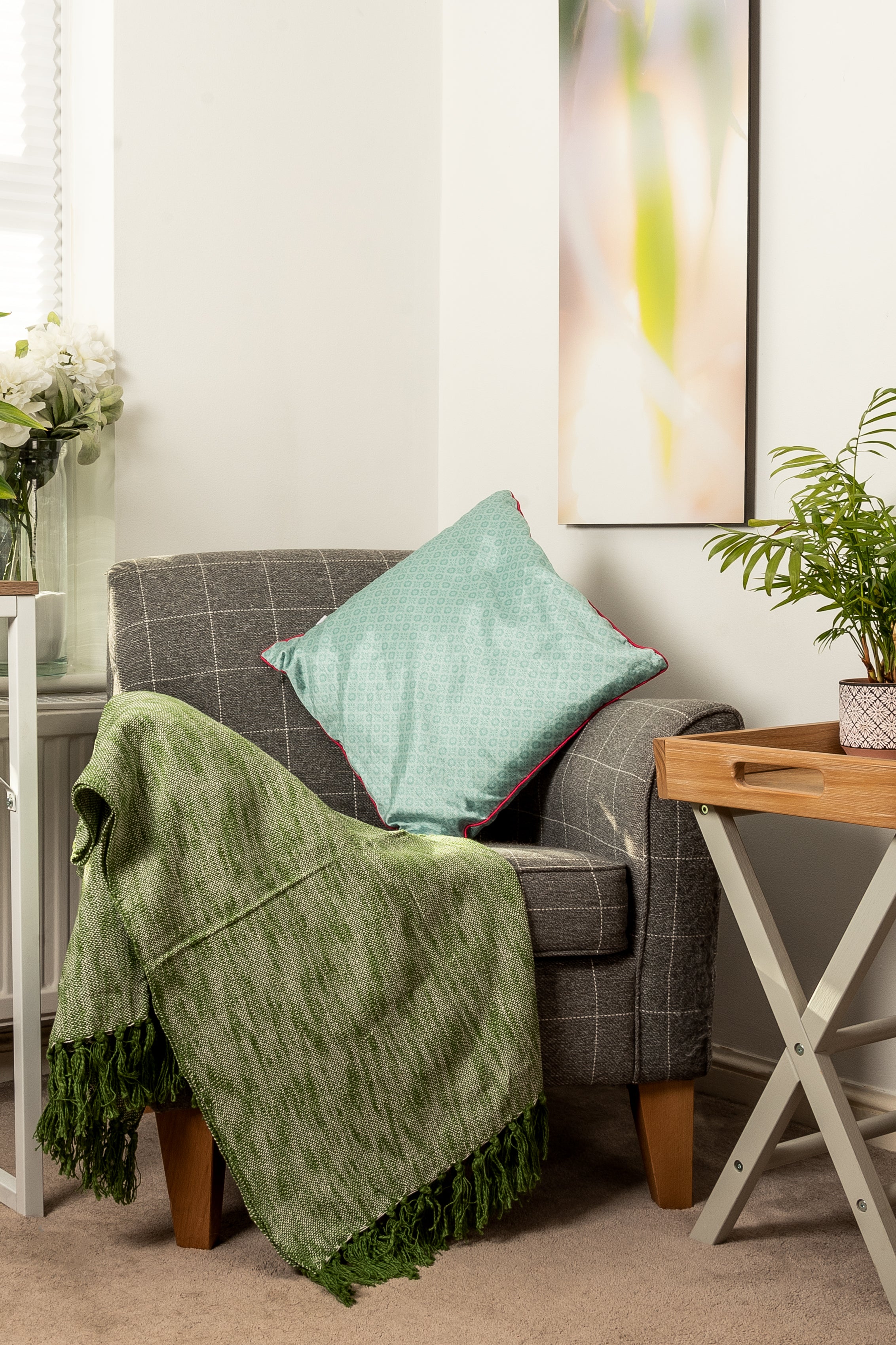 A cozy corner featuring a grey patterned armchair with a green textured throw blanket and a teal cushion. To the right is a small wooden side table with a plant, and a tall, vertical abstract painting hangs on the wall behind it.