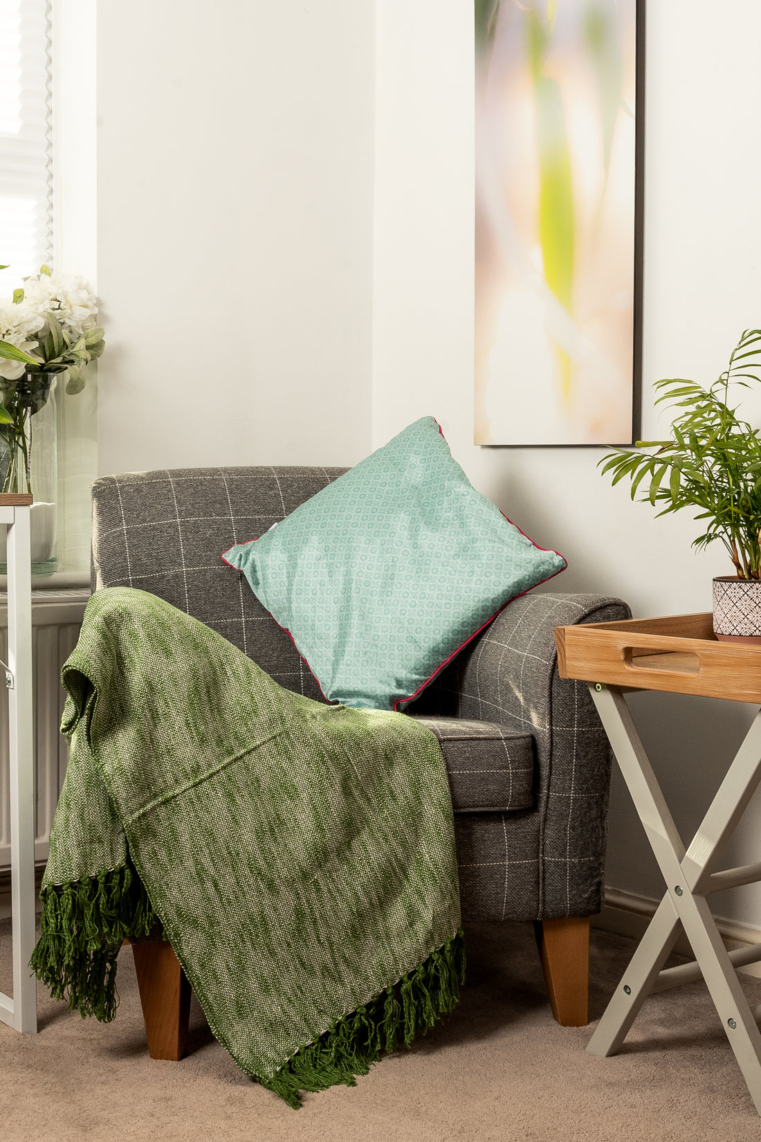 A cozy corner featuring a grey patterned armchair with a green textured throw blanket and a teal cushion. To the right is a small wooden side table with a plant, and a tall, vertical abstract painting hangs on the wall behind it.
