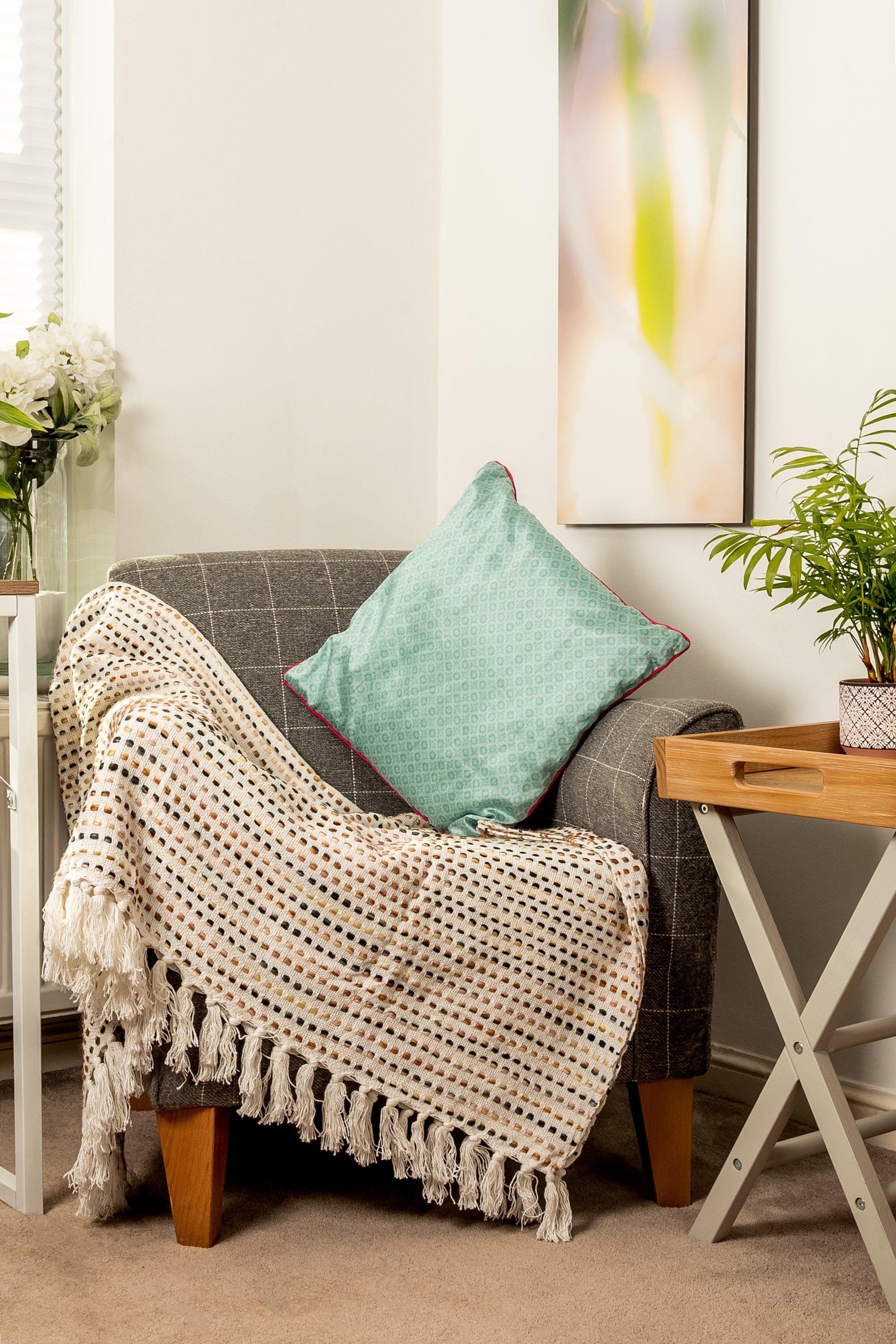 A cozy reading nook featuring a grey armchair draped with a cream and black patterned fringed throw and a teal cushion. To the right is a wooden tray on an X-frame side table, set against a wall with a tall abstract painting.