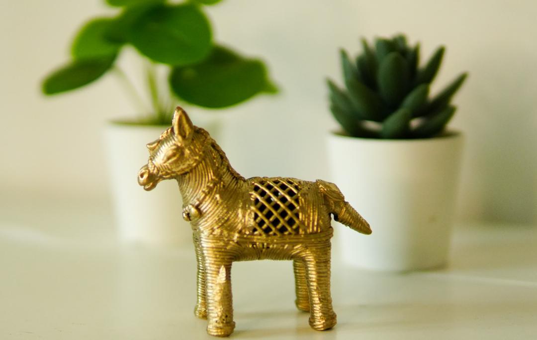 A gold-colored Dhokra horse figurine statue placed on a surface with two potted plants in the background.