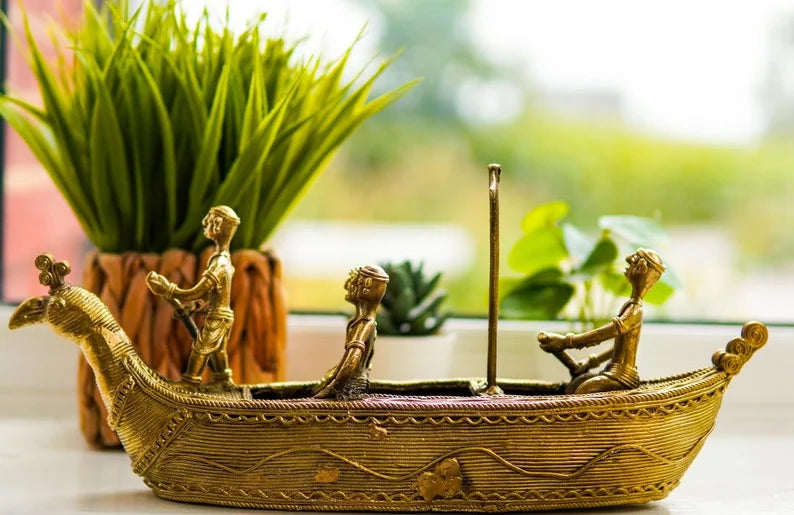 This item is a handcrafted Dokra brass boat showpiece featuring tribal figures, often used for home decor. 