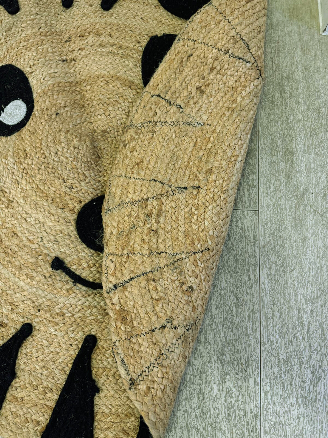 A woven straw or jute rug partially folded on a light wooden floor, featuring a playful face design with black eyes and a smiling mouth, showing the braided texture and stitching on the underside.
